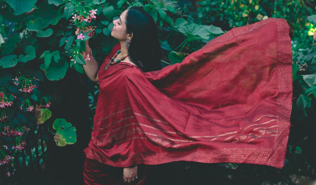Woman in a flowing red saree stands among lush green foliage, embracing traditional Indian beauty.