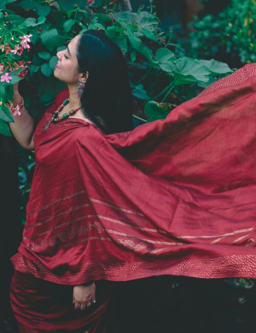 Woman in a flowing red saree stands among lush green foliage, embracing traditional Indian beauty.