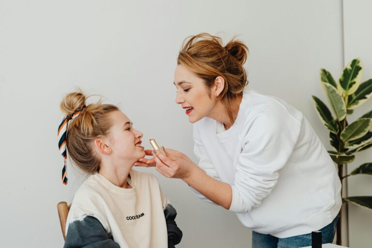 A mother lovingly applies lipstick to her smiling daughter indoors, capturing a tender family bonding moment.